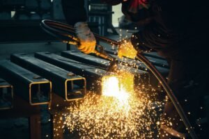 A welder sparks metal beams indoors in an industrial factory setting.